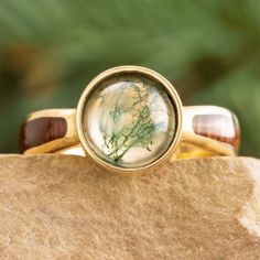 This one-of-a-kind ring features a stunning and distinctive moss agate cabochon, forming a beautiful hybrid stone. Perfect for everyday wear or as a wedding ring, the solid yellow gold bezel setting perfectly highlights the large cabochon stone. The ring's tapered shank is expertly adorned with ironwood, adding a natural and striking element to the design. Create the perfect bridal set by pairing this ring with one of our unique, custom women's wedding bands. RING LAYOUT Ring Width: 3.5 mm Ring Gold Agate Cabochon Rings, Adjustable Moss Agate Artisan Jewelry, Nickel-free Bohemian Moss Agate Jewelry, Unique Cabochon Moss Agate Jewelry, Bezel Set Cabochon, Women's Wedding Bands, Nature-inspired Moss Agate Jewelry With Natural Inclusions, Handcrafted Engagement Ring, Moss Agate Engagement Ring