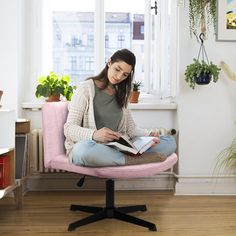 a woman sitting on a pink chair reading a book in her living room with potted plants