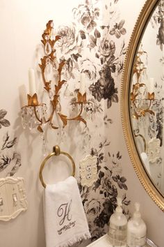 a bathroom with floral wallpaper and gold accents on the mirror, toilet paper towel holder, soap dispenser and candle holders