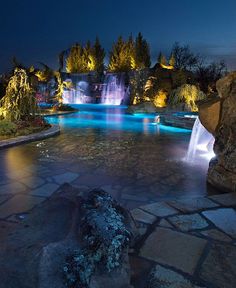 an outdoor pool at night with lights on the water and waterfall in the middle, surrounded by rocks