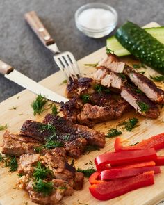 a wooden cutting board topped with meat and veggies next to a knife and fork