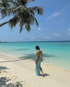 a woman in a blue dress walking on the beach next to a palm tree and water