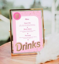 a pink and gold drink menu sitting on top of a wooden table next to a white feather