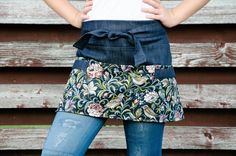 a woman wearing an apron and jeans with her hands on her hips, standing in front of a wooden wall