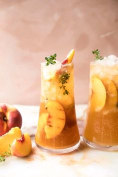 two glasses filled with ice and fruit on top of a marble table next to peaches