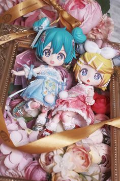 two dolls are sitting in a box with flowers around them and ribbons on the sides