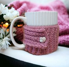 a knitted coffee mug cozys up next to some flowers and a pink blanket