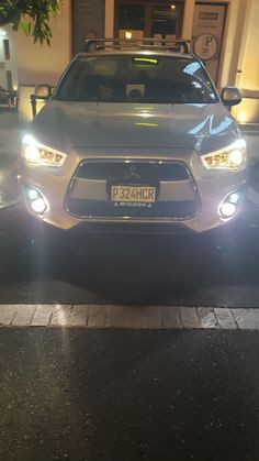 the front end of a silver car parked in front of a building with lights on