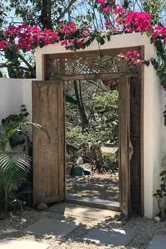 an open wooden door with pink flowers growing on the wall and behind it is a stone walkway