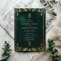 a green and gold wedding card on top of a marble table next to greenery