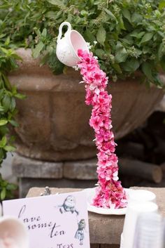 pink flowers are growing out of a white teapot on a table next to a sign