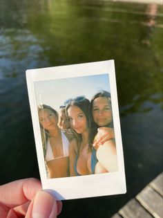 a person holding up a polaroid with three girls in front of the camera and water behind them