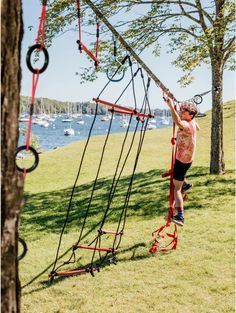 a young boy is climbing on a rope course in the park with boats in the water behind him