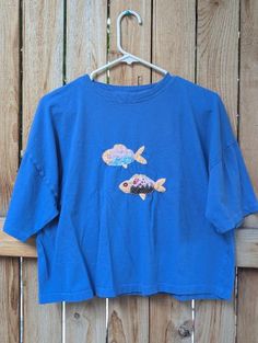 a blue t - shirt with two fish on it hanging from a wooden fence,