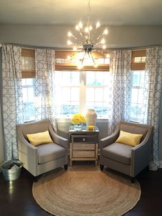 a living room with two chairs and a table in front of a window that has yellow pillows on it