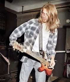 a man with long hair playing an electric guitar in a recording studio while wearing jeans and a checkered shirt