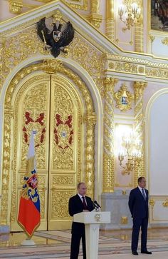 two men in suits are standing at a podium with flags and an eagle on the wall behind them