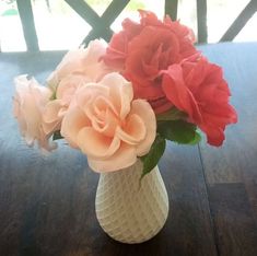 a white vase filled with pink and red flowers on top of a wooden table next to a window