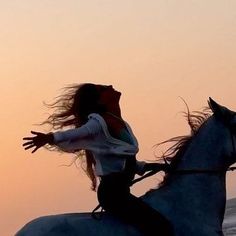 a woman riding on the back of a white horse next to the ocean at sunset