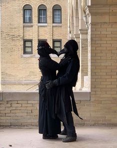 two people dressed in black standing next to each other near a brick building with arched windows