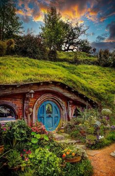 the hobbot house is surrounded by plants and flowers in front of a cloudy sky