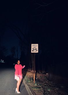 a woman in a pink dress standing on the side of a road at night with a speed limit sign