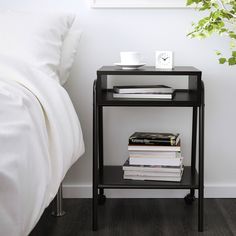 a black end table with some books on it next to a white bed and plant