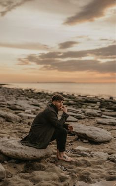 a man sitting on top of a rock covered beach next to the ocean while talking on a cell phone