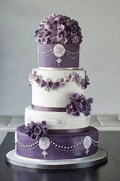 a three tiered cake with purple flowers and pearls on the top, sitting on a table