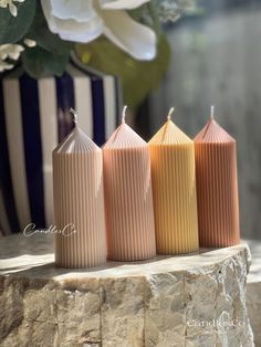 three different colored candles sitting next to each other on a stone slab with flowers in the background