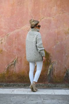 a woman standing in front of a pink wall wearing white jeans and a gray sweater