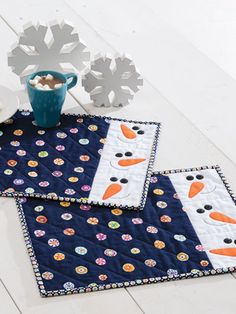 two quilted placemats with snowmen on them and a cup of hot chocolate