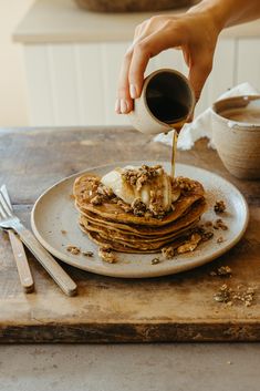 a person pouring syrup onto a stack of pancakes on a plate with butter and granola