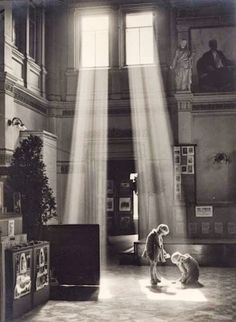 an old black and white photo of two people in a room with sunlight streaming through the windows