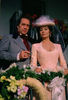 two people standing next to each other in front of flowers and wreaths on a balcony