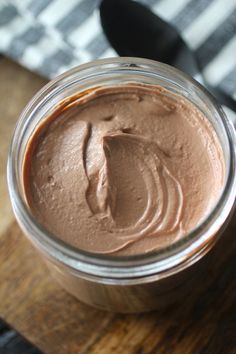 a jar filled with chocolate frosting sitting on top of a wooden table next to a spoon