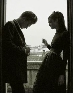 two people standing next to each other in front of a window looking at their cell phones