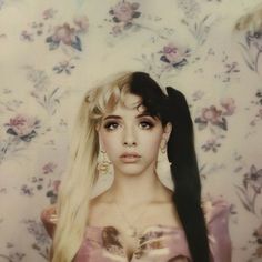 a woman with long blonde hair standing in front of a floral wallpapered background