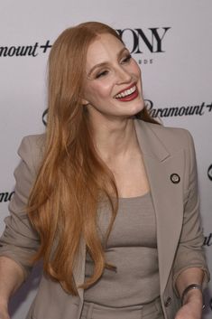 a woman with long red hair smiling and wearing a blazer over a gray dress