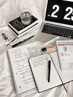 an open laptop computer sitting on top of a bed next to notebooks and pens