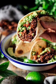 a hand holding a burrito over a bowl filled with salsa and guacamole