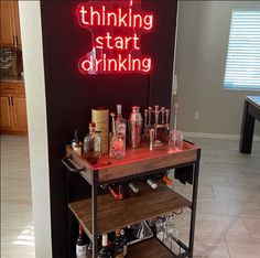 a bar cart with liquor bottles and glasses on it in front of a neon sign that says thinking start drinking