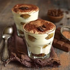 two glasses filled with dessert sitting on top of a wooden table next to spoons