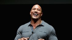 the rock smiling while standing on stage with his hands out and wearing a gray shirt