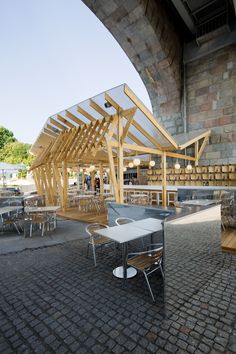 an outdoor seating area with tables and chairs under a wooden structure on the side of a brick wall