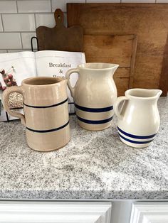 three white and blue pitchers sitting on top of a counter next to a cookbook