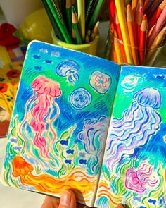 someone holding up an art journal with colored pencils in front of them and jellyfish drawings on the pages