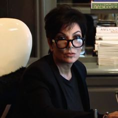 a woman with glasses sitting at a desk in front of a lamp and bookshelf