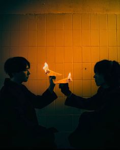 two people sitting in front of a yellow wall holding small lights up to their faces