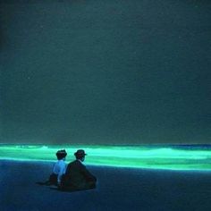 two people are sitting on the beach looking at the water with green lights in the background